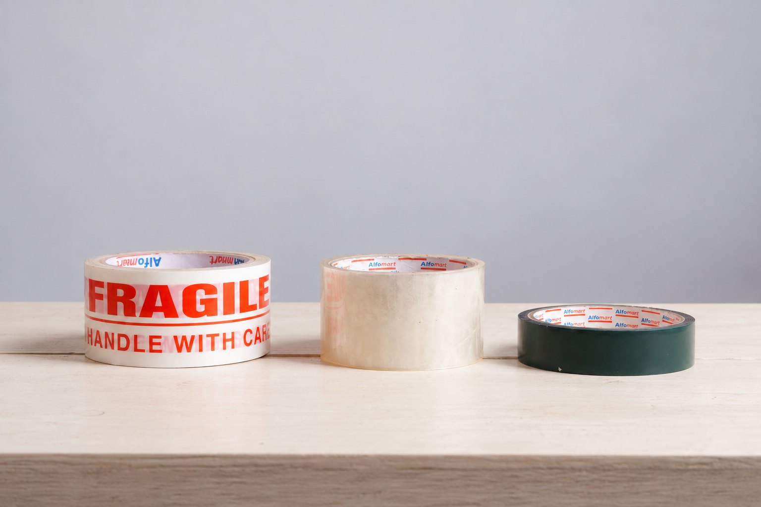 Wide printed and clear tape rolls beside a narrower dark green roll on a wooden surface